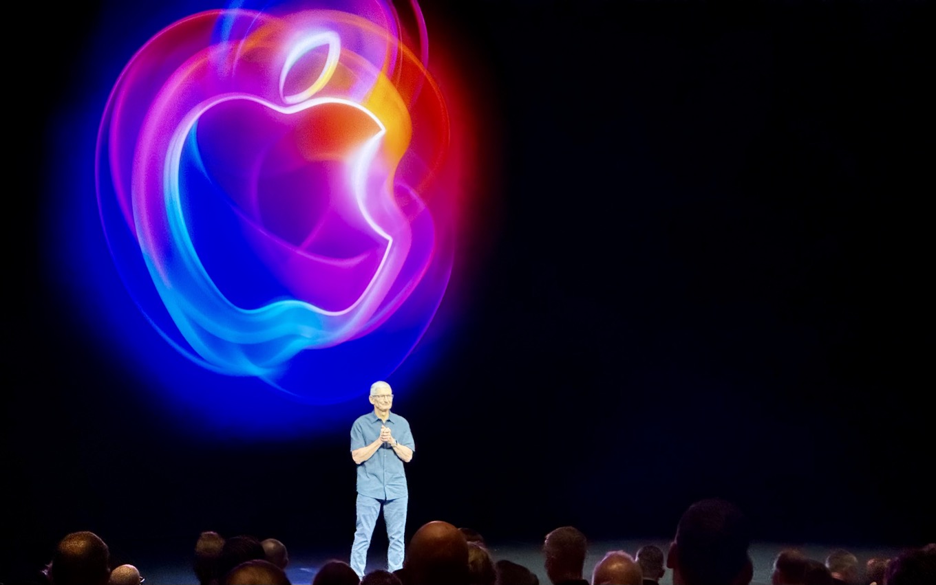 Tim Cook standing on a dark stage with a large, colorful Apple logo projected behind him on stage screens