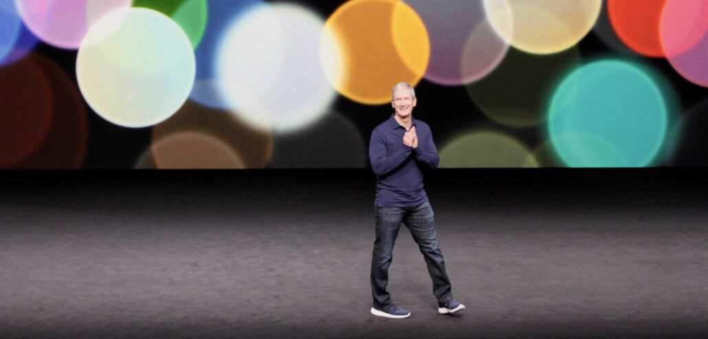 Man standing on a dark stage wearing a navy sweater and jeans, with a large colorful bokeh backdrop behind him.