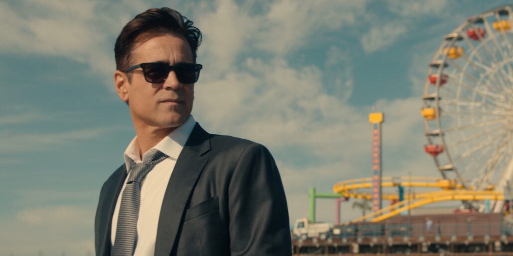 Apple TV unveils return of hit detective series with Colin Farrell - Dans Tutorials - Dans Tutorials Man in a dark suit and sunglasses stands at an amusement park with a ferris wheel in the background on a sunny day.