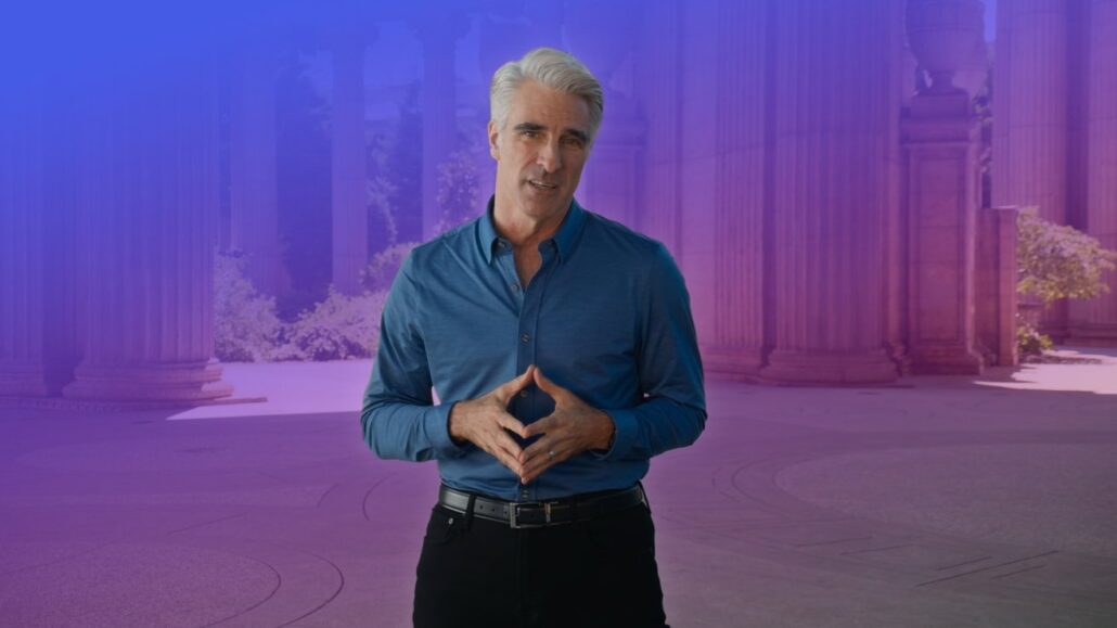 Adult man in a blue shirt stands with hands together, speaking in front of a purple-toned columned backdrop.