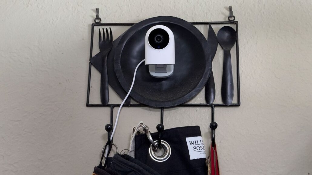 White security camera mounted on a black metal wall decor plate over a utensil-hanging rack with bags below.