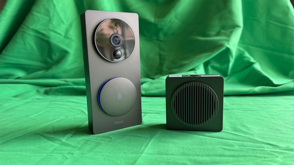 Smart doorbell with circular camera housing and a secondary round touch/interface on a gray panel, standing upright next to a black square speaker unit on a green backdrop.