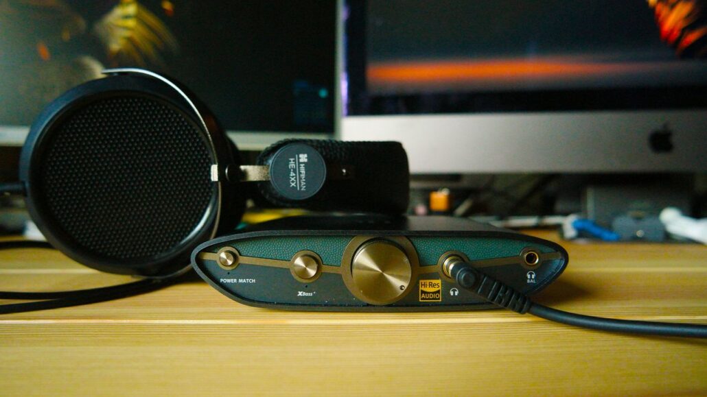 Over-ear headphones rested on a wooden desk, plugged into a small DAC/amp with a large gold volume knob and a Hi-Res Audio label. A computer monitor sits in the background.