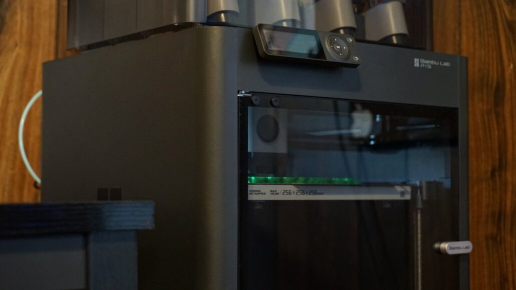Bambu Lab 3D printer in a dark enclosure with a glass front and a control display mounted on top against a wooden wall.