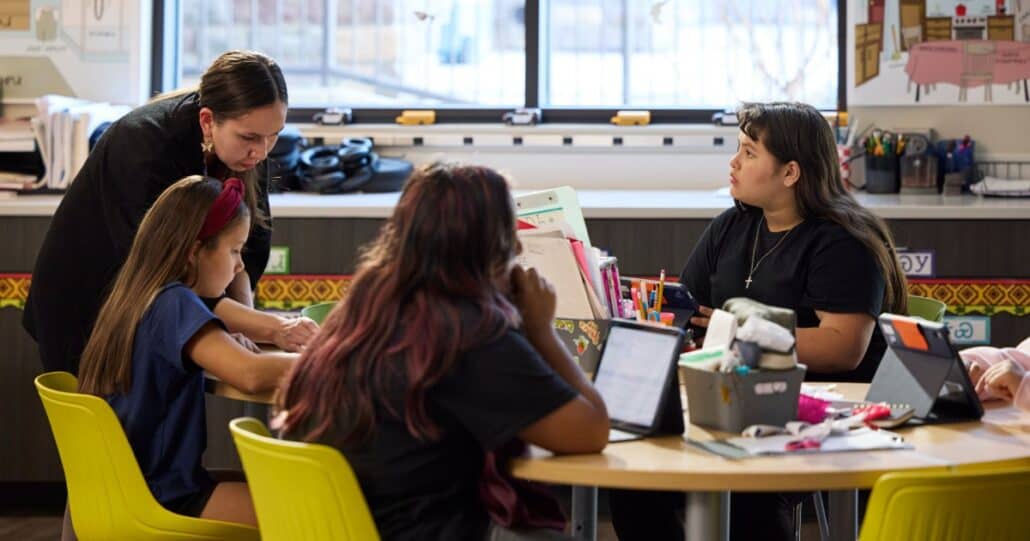 Teacher assists two students at a round table in a classroom, with yellow chairs and tablets on the table.