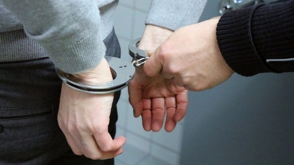 Crime blotter Man arrrested for 000 iPhone theft - Dans Tutorials - Dans Tutorials A person with wrists in metallic handcuffs being restrained by another hand from behind a counter or table edge.