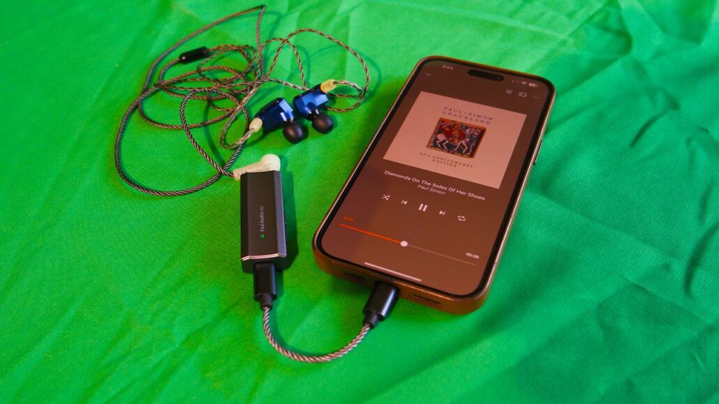 Phone playing Paul Simon's Graceland on a green surface, connected to braided wired earphones and a small inline amplifier/dongle beside it.