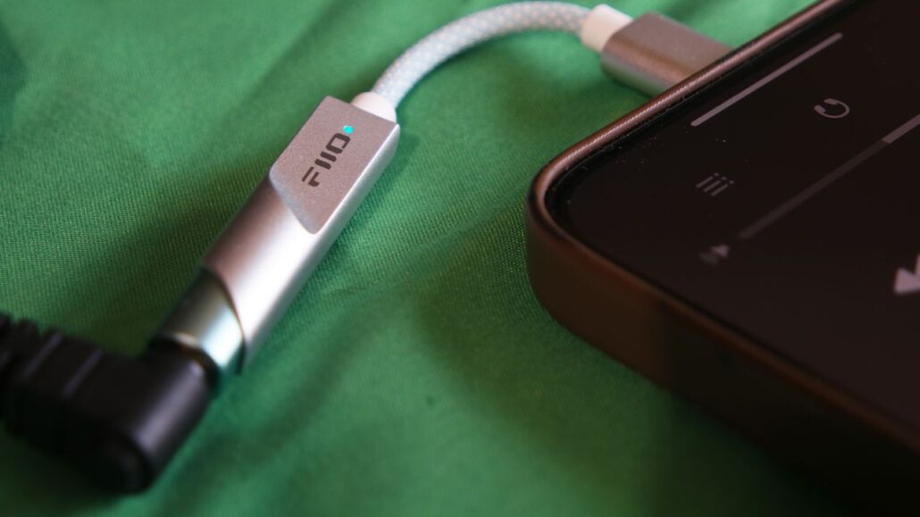 Close-up of a silver FiiO connector on a braided cable resting beside a smartphone on green fabric, showing a blue indicator light on the connector.
