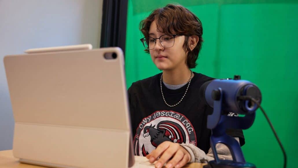 How students and teachers are using iPad to save an endangered language - Dans Tutorials - Dans Tutorials Person with short hair and glasses sits at a desk using a tablet, with a blue microphone on the right and a green screen behind them.