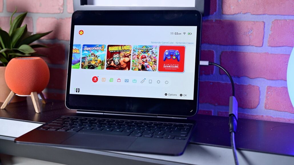 Tablet with a keyboard dock displaying a row of game icons on a desk against a brick wall. A small orange speaker sits to the left.