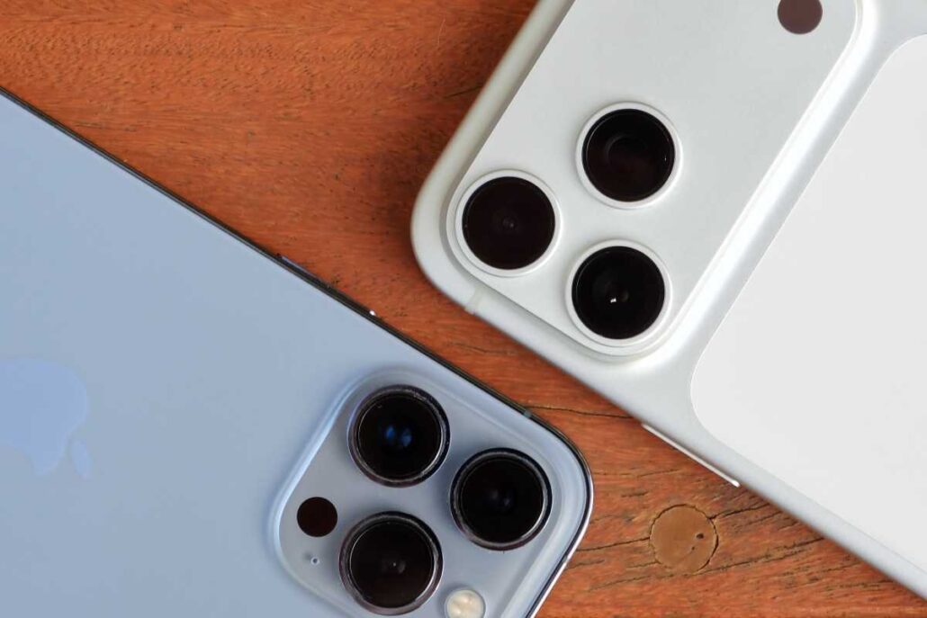 Two iPhones with triple-camera setups (blue and white) resting on a wooden surface.
