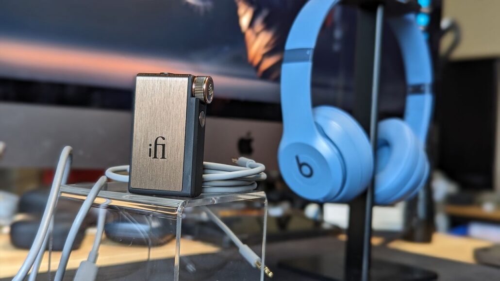 Small brushed-metal iFi audio device on a clear stand with white cables, blue over-ear headphones in the background on a desk.