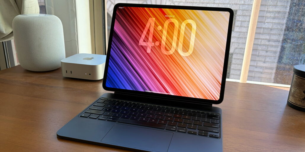 Laptop with colorful diagonal gradient wallpaper on a wooden desk, flanked by a white HomePod and a silver Mac mini.