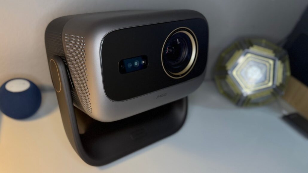 Close-up of a compact projector on a curved stand, with its lens facing forward and side ventilation visible.