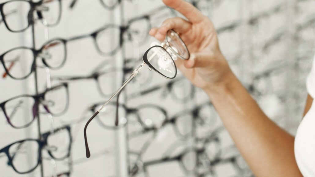 Like Apple Watch at start Apples smart glasses plan will challenge the entire industry - Dans Tutorials - Dans Tutorials Person holds a pair of rimmed glasses, with many frames displayed on the wall behind in an optical shop.