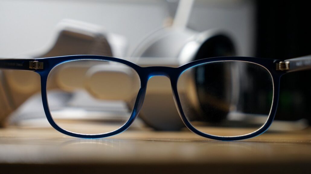 Longtime leaker joins others in saying Apple Glasses wont arrive until late 2027 - Dans Tutorials - Dans Tutorials Blue-framed eyeglasses resting on a wooden desk with blurred headphones in the background.