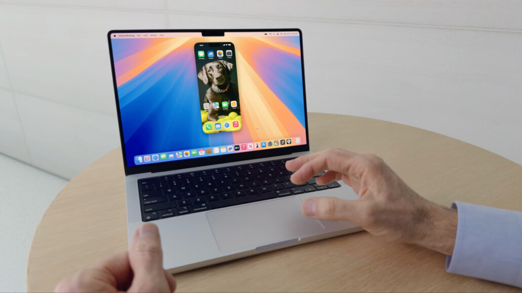 Laptop on a round wooden table showing colorful wallpaper with an iPhone-like screen overlay; a person’s hands hover near the keyboard and trackpad.