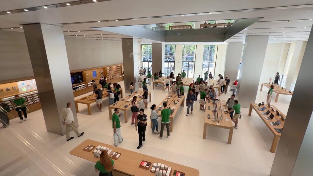 Open electronics store with wooden tables displaying laptops; customers browse as staff in green shirts assistingshopping.