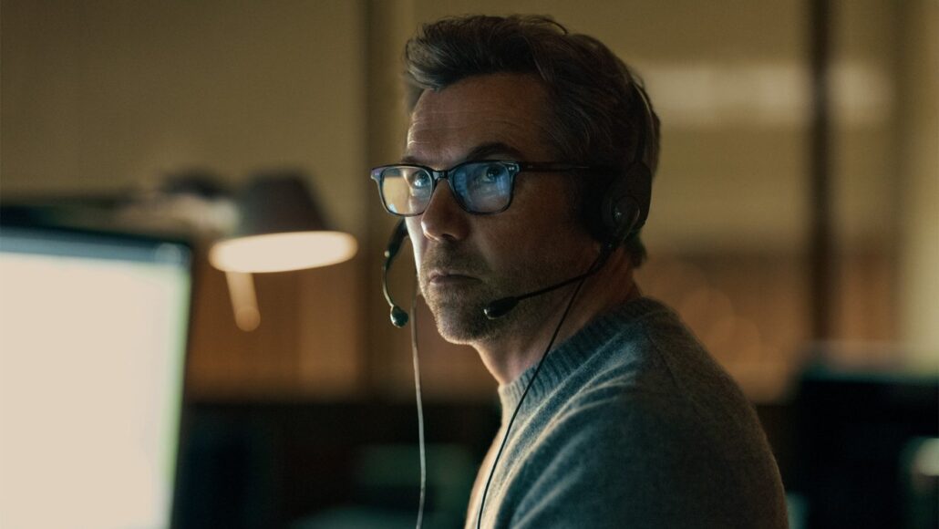 Middle-aged man wearing glasses and a headset with microphone, looking at a computer screen in a dim office to assist a caller on a support line
