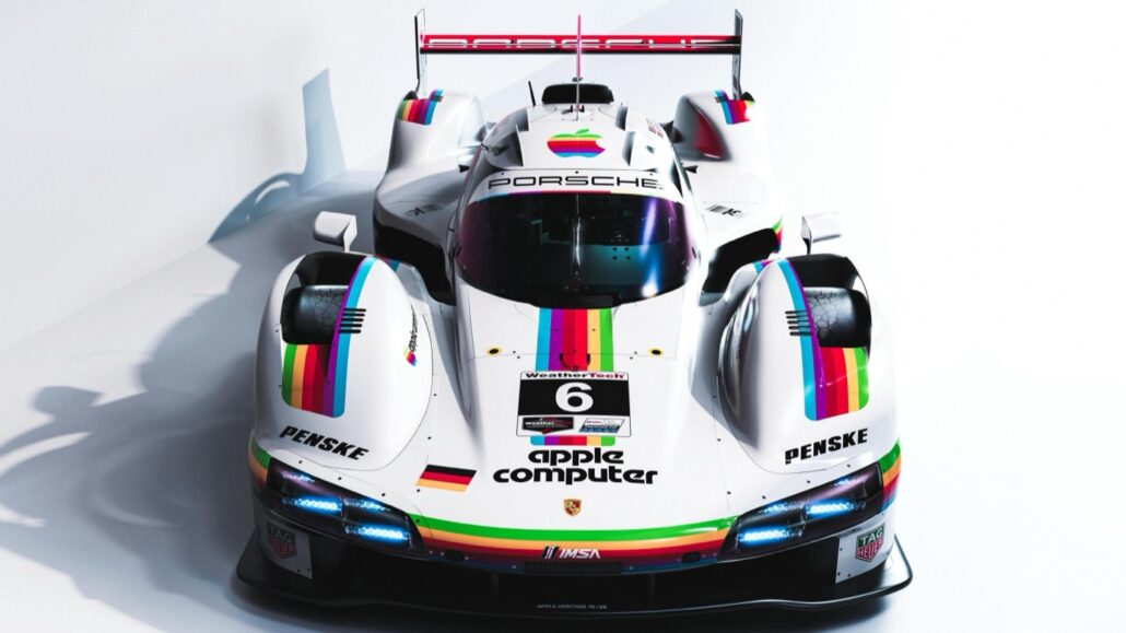 White Porsche race car with rainbow stripe livery and sponsor logos, viewed from the front center. Reads 'Porsche' on the windshield area and features blue headlights at the front." ,