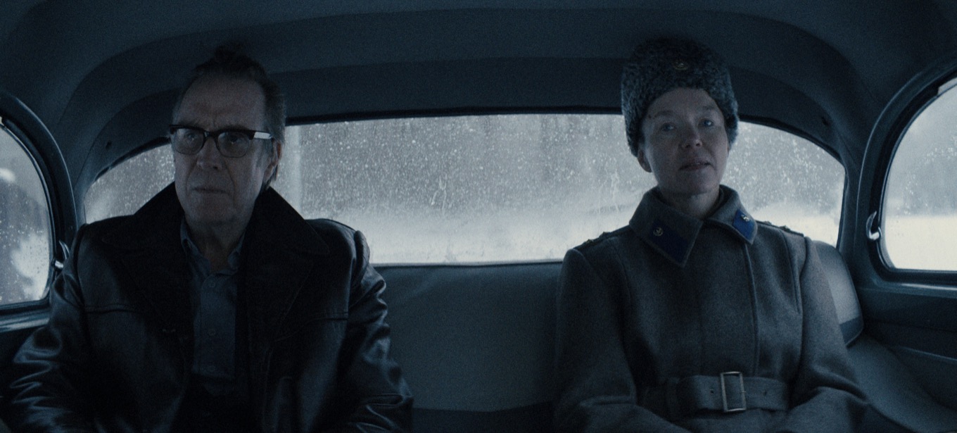 Two people sit in the backseat of a car wearing heavy coats on a snowy day, windows frosted.