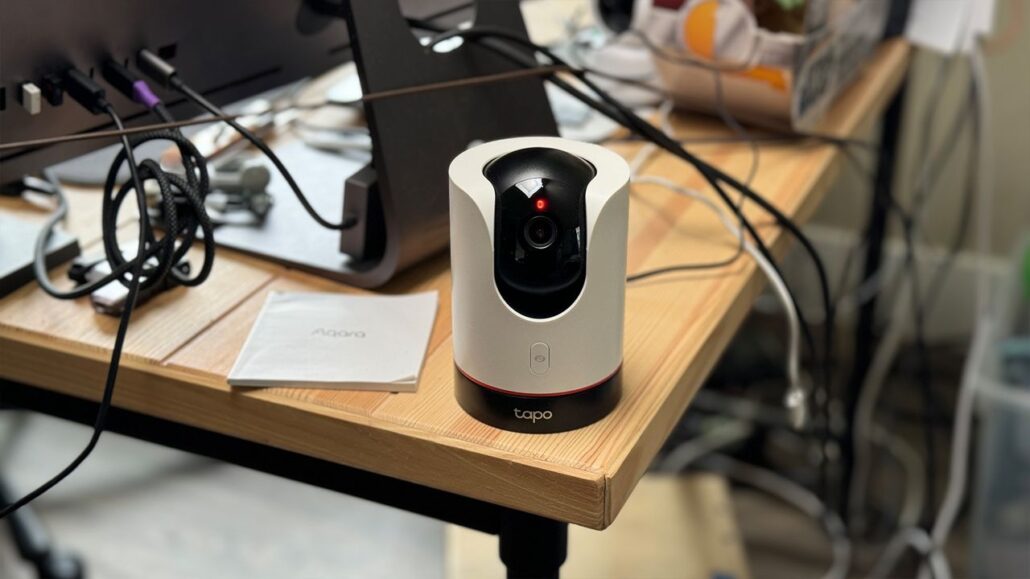 White cylindrical webcam on a wooden desk with tangled cables and a red indicator light at the front (Tapo camera).
