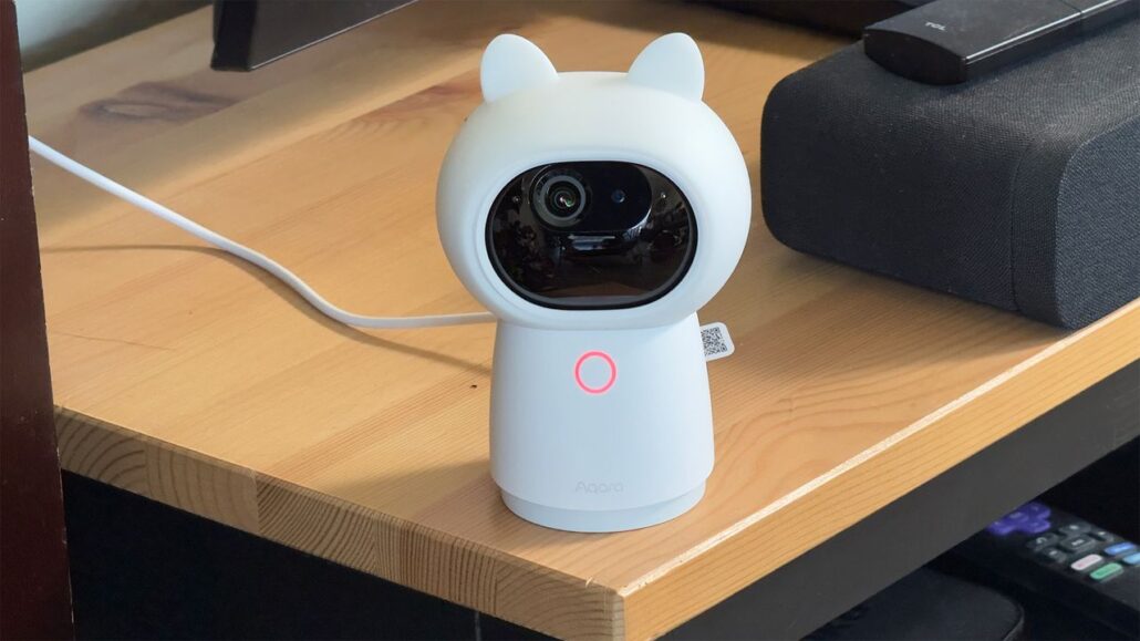 White cat-ear shaped security camera on a wooden desk, lens centered and pink ring LED visible.