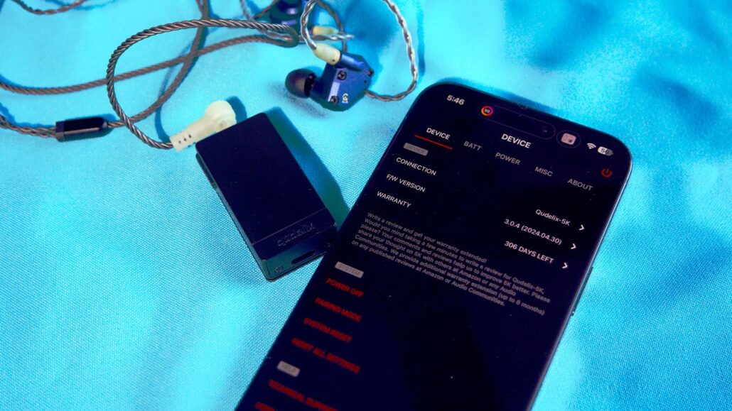 Smartphone on a bright blue fabric displaying device settings screen beside a small rectangular black audio adapter with braided cables and in-ear monitors nearby