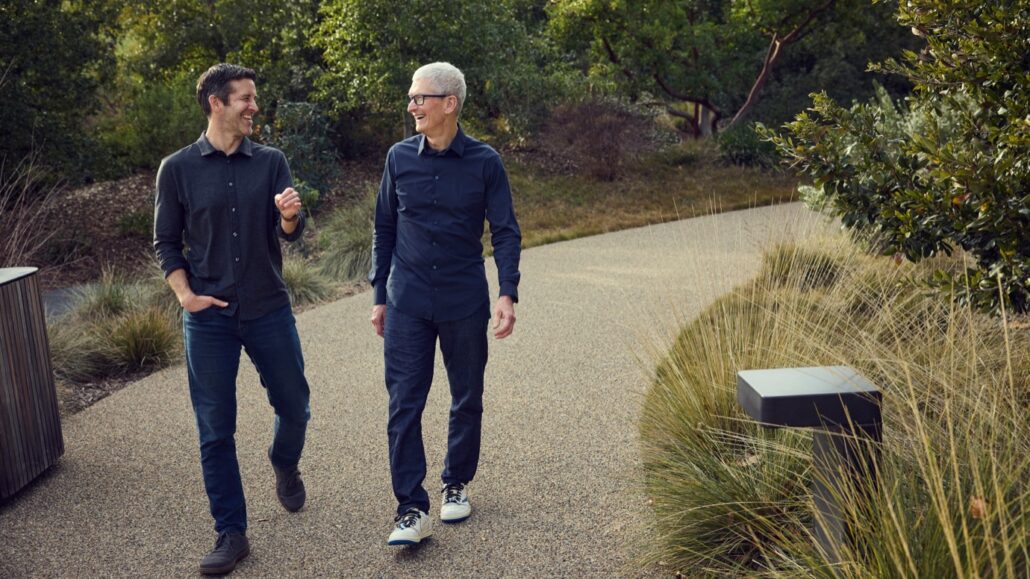 Two men walk and chat along a park path surrounded by shrubs and grasses. They wear casual dark shirts and jeans, enjoying a sunny outdoor stroll.