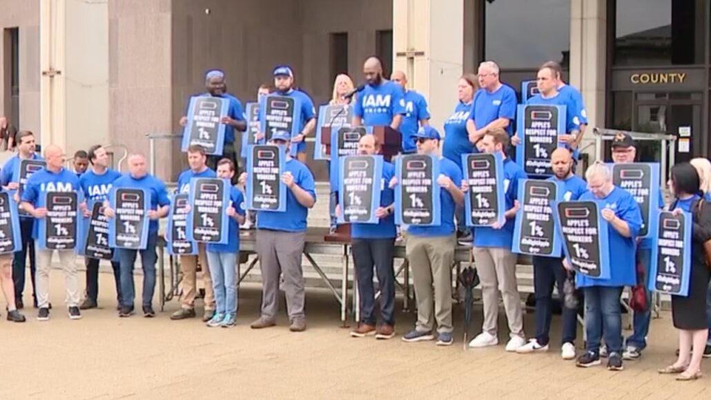 Group of people in blue shirts standing on a plaza in front of a county building, each holding tall protest signs.