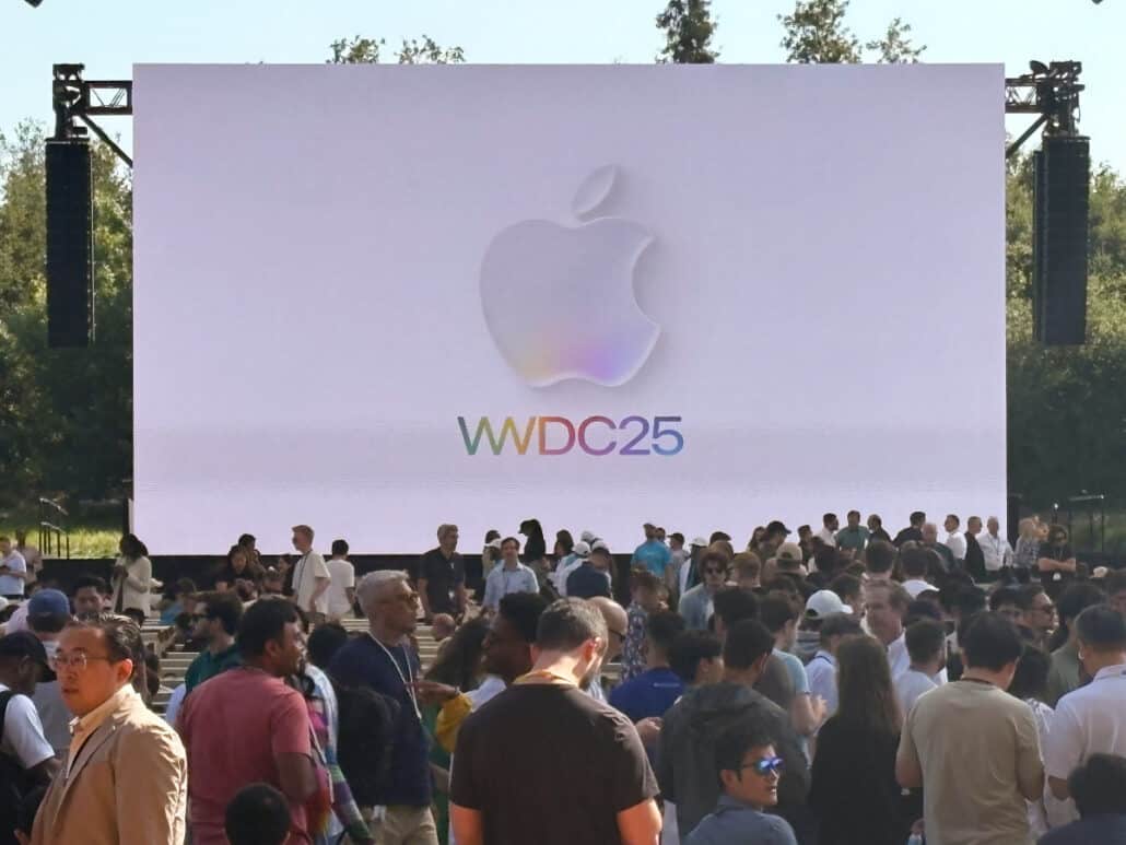 Outdoor Apple event: large screen with Apple logo and 'WWDC25' above a crowded audience of attendees.