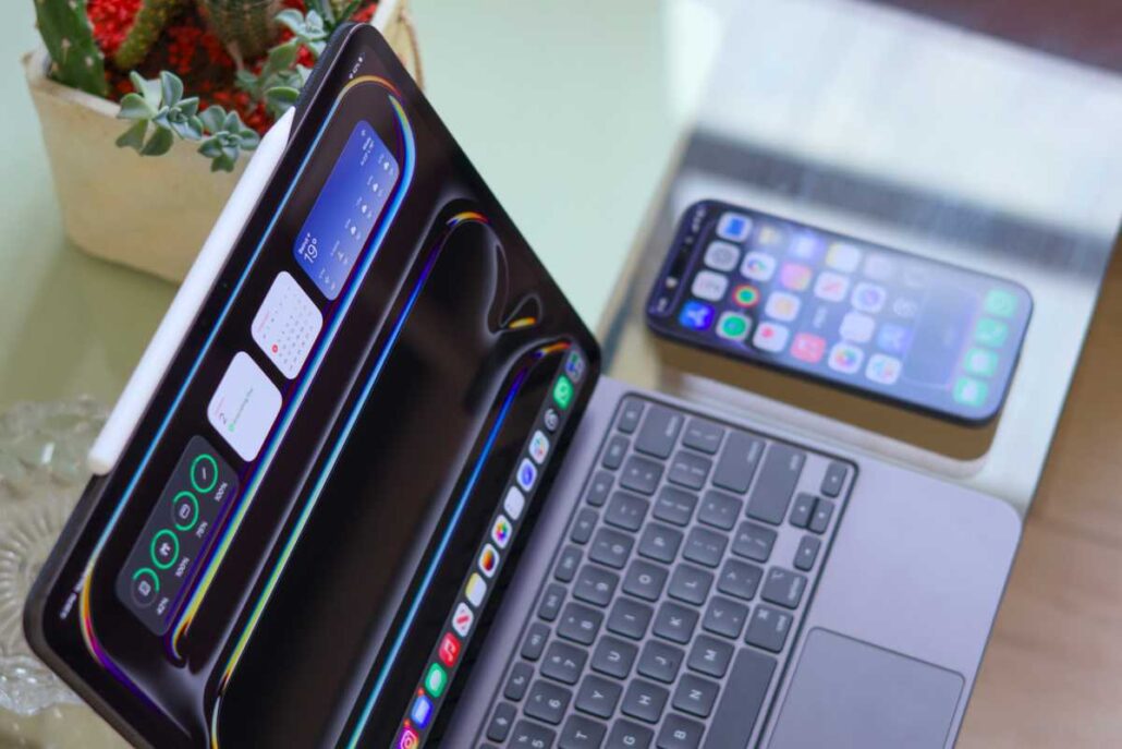 Workspace with a tablet (iPad Pro) leaning on its side, a laptop keyboard visible, and an iPhone on a stand nearby on the desk.