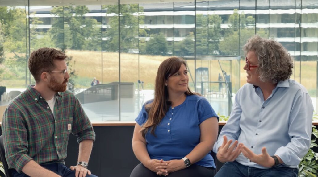 Three coworkers seated in a glass-walled, sunlit office, engaged in a discussion.
