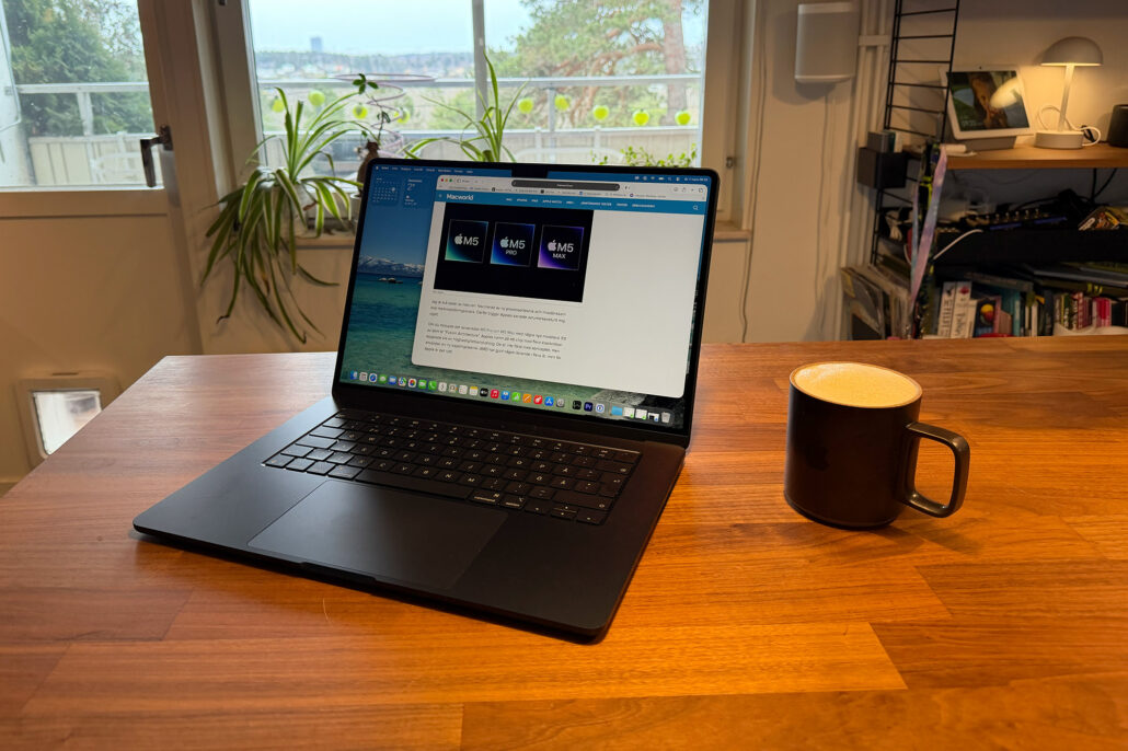 Laptop on a wooden desk with a black mug to the right, greenery and a window in the background.