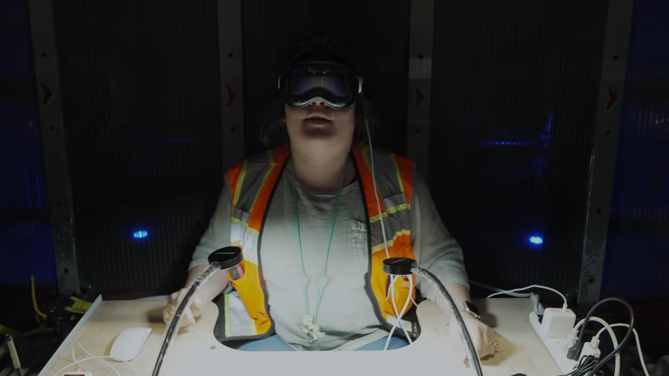 Person wearing a safety vest and virtual reality headset sits at a desk with cables in a dark room.