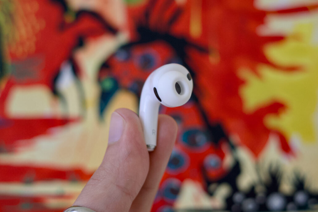 Apples AirPods Pro 3 crash to an all-time low price of 9 - Dans Tutorials - Dans Tutorials Close-up of a white wireless earbud held between fingers against a colorful blurred red-yellow background, showing the speaker mesh.