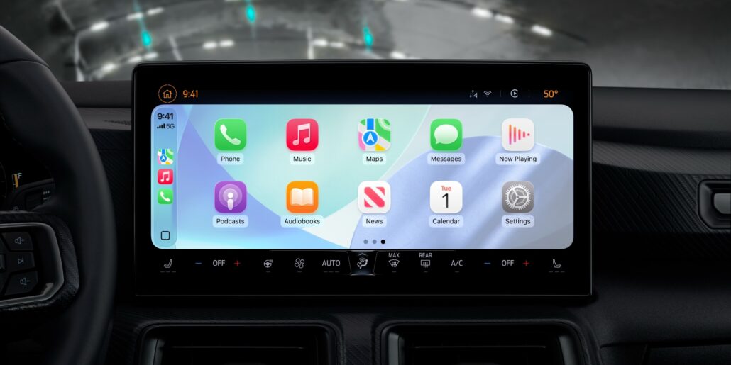 Car dashboard touchscreen displaying an iPhone-style home screen with apps like Phone, Music, Maps, Messages, Now Playing, Podcasts, Audiobooks, News, Calendar, and Settings