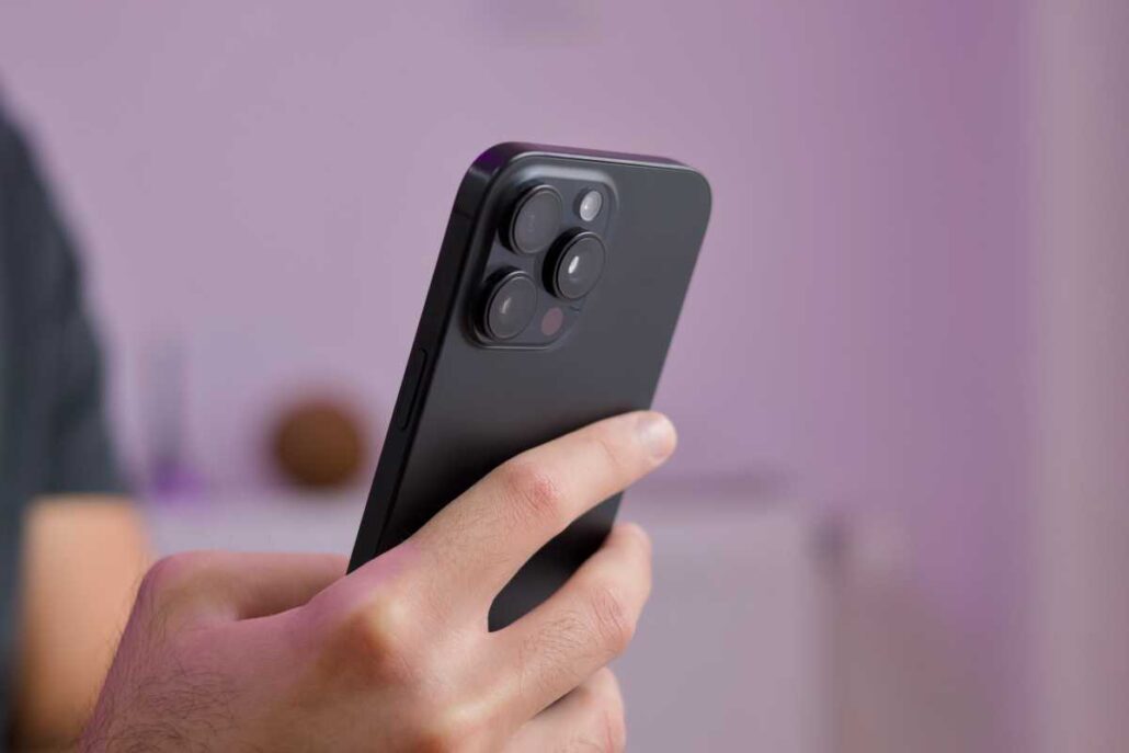 Close-up of a hand holding a dark smartphone from the back, showing a multi-lens camera module against a purple background.