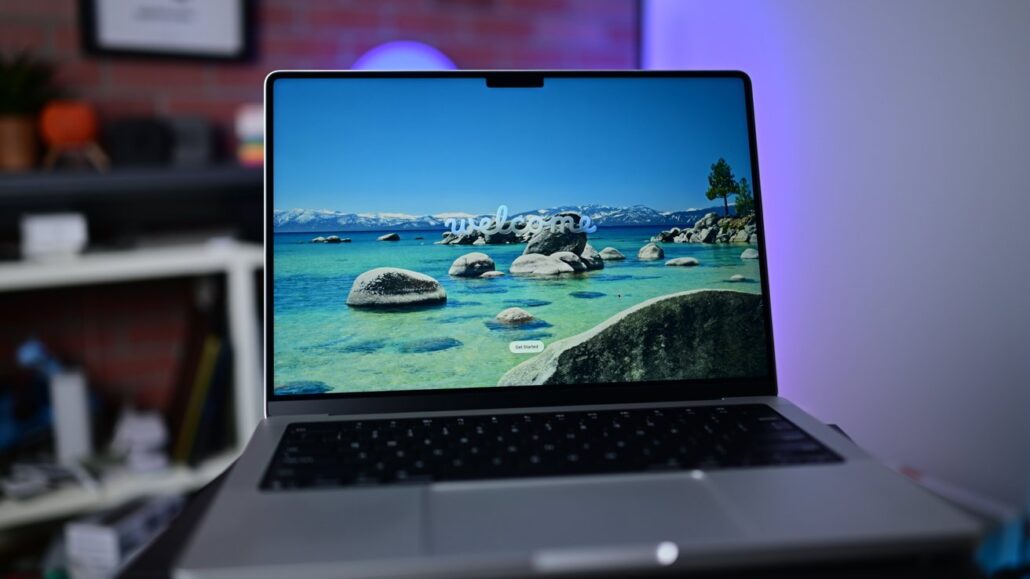 Open laptop on a desk showing a tropical lake wallpaper with rocks, clear water, and a small 'welcome' text on screen (room blurred in background).