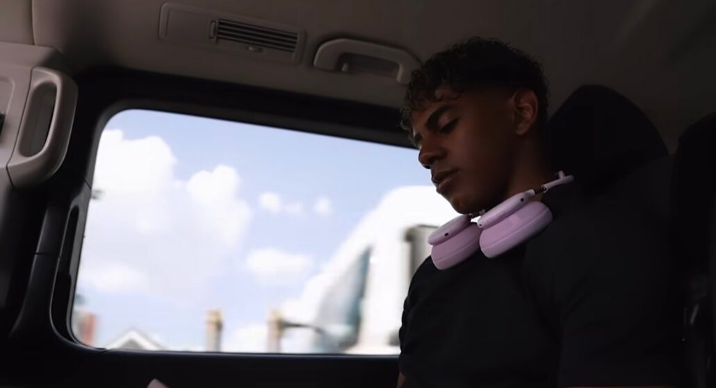 Young man wearing pink over-ear headphones sits in a car, looking down, with a blue sky and clouds outside the window.