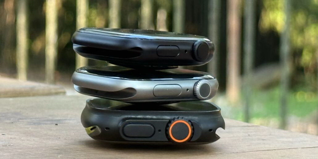 Heres how Apple Watch Series 11 stacks up against Oura Fitbit and Whoop per WSJ - Dans Tutorials - Dans Tutorials Stacked smartwatches on a wooden table outdoors; black top, silver middle, and dark bottom with an orange ring on the bezel.
