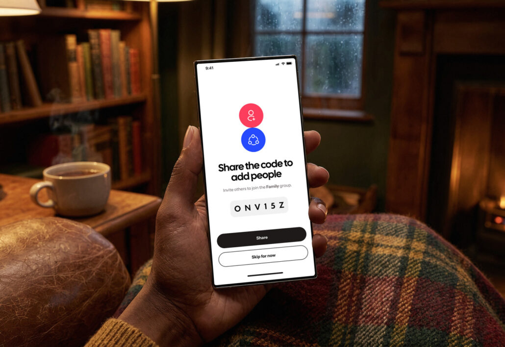 HeyPolo is a smarter Find My app for family friends and co-workers - Dans Tutorials - Dans Tutorials Person holding a smartphone showing a 'Share the code to add people' screen indoors by a fireplace and mug on a wooden table to invite others to a family group.
