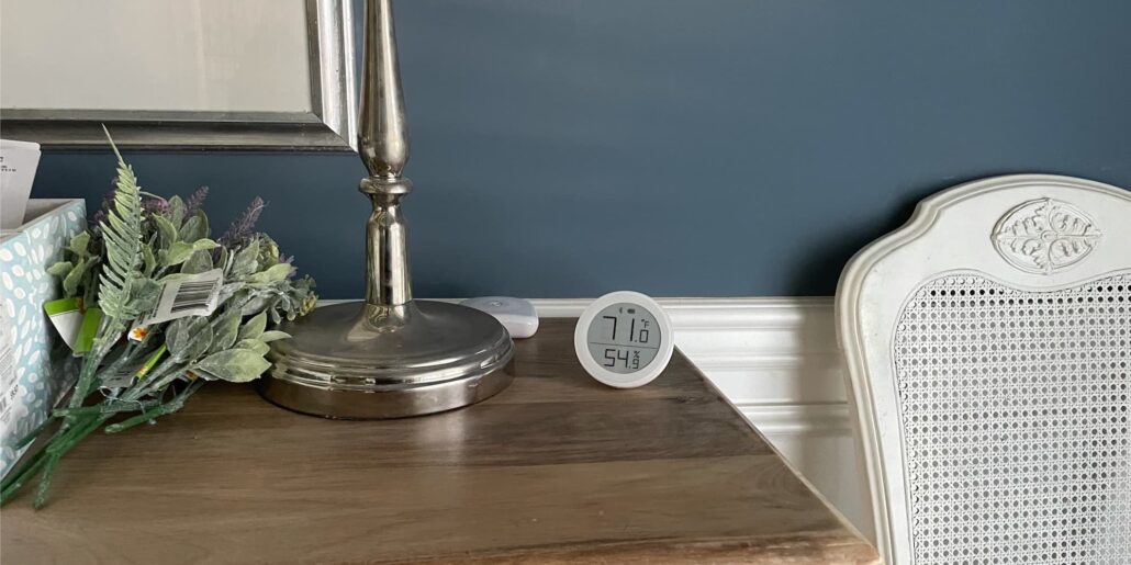 Silver lamp on a wooden table with a bouquet of faux greenery and a round digital gauge showing 71°F and 54% humidity against a blue wall.