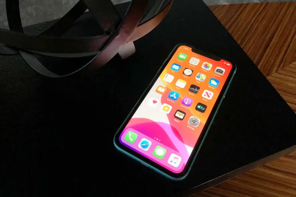 iOS 27 Everything we know about the 2026 iPhone update - Dans Tutorials - Dans Tutorials Green iPhone with colorful home screen on a black desk, near a decorative metal sculpture.