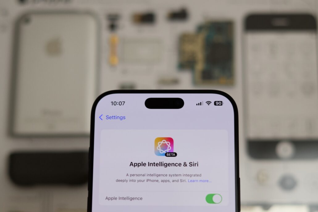 Close-up of an iPhone screen showing the Settings page for Apple Intelligence & Siri, with a green toggle for Apple Intelligence on and blurred background devices behind.