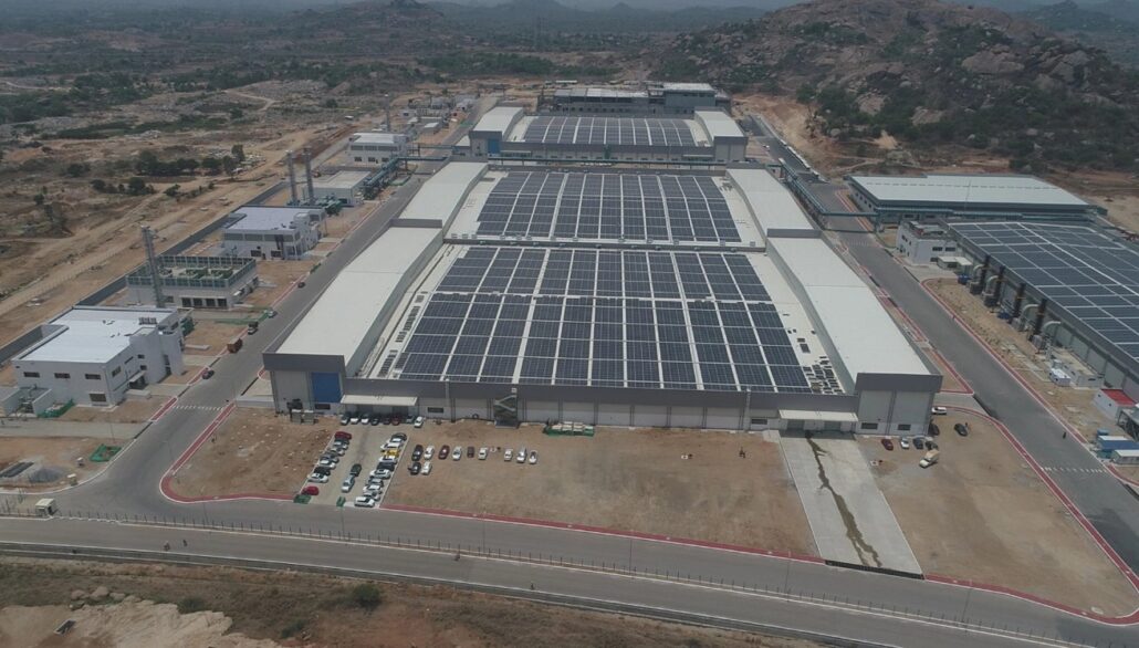 Aerial view of a large solar power plant with rows of PV panels covering long white-roofed industrial buildings in a desert landscape.