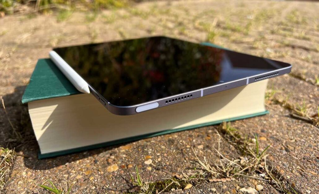 Tablet resting on a thick hardcover book outdoors on a dirt surface, with a white stylus beside it.