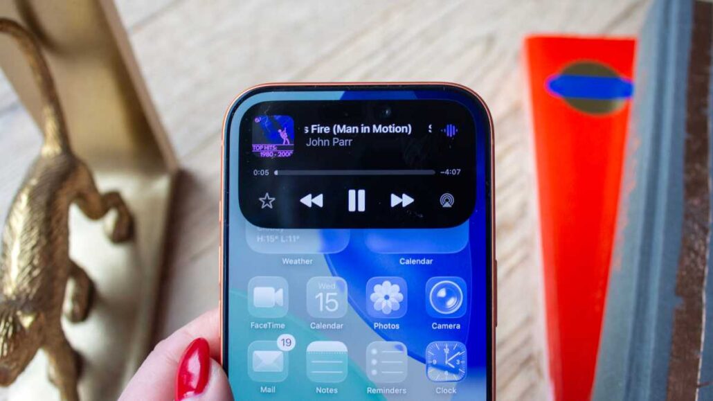 iPhone screen displaying a music player playing John Parr’s 'Fire (Man in Motion)' with app icons (Photos, Calendar, Weather) visible below, held by a person with red-painted nails.