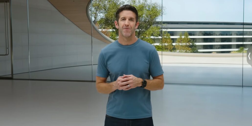 Man in a blue T-shirt stands in a bright, modern glass-walled space, hands clasped as he speaks to the camera.