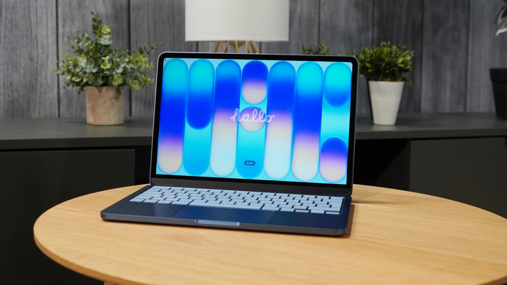 Laptop with blue and cyan abstract wallpaper on a round wooden table in a modern office, with potted plants in the background.